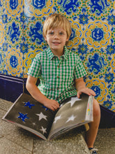 Load image into Gallery viewer, Child in a green checkered shirt sitting on the floor with a book against a colorful tiled wall.