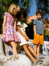 Load image into Gallery viewer, Three children sitting on a decorative bench outdoors with trees and a clear sky in the background.