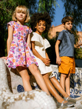 Load image into Gallery viewer, Three children sitting on a decorative bench outdoors with trees and buildings in the background.