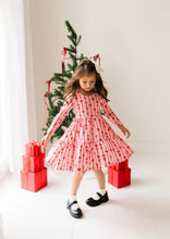 Load image into Gallery viewer, Child looking down while twirling in the pink Dainty Bows dress, showing its full skirt and red bow-and-stripe print.