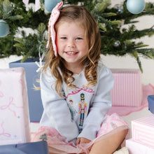 Load image into Gallery viewer, A young girl with a pink bow in her hair sits under a decorated Christmas tree, wearing a light blue knit sweater with a nutcracker graphic and a pink tulle skirt. Gift boxes are visible around her.