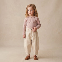 Load image into Gallery viewer, A toddler girl stands against a warm beige textured studio backdrop, wearing a dusty mauve long-sleeve top with a ruffled collar, cream-colored pleated trousers, and brown leather shoes.