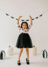 Load image into Gallery viewer, A young girl with dark hair wearing a pink long-sleeve top with a black cat pattern and a black tulle tutu skirt, posing with her arms raised in a playful Halloween pose. She is standing in a bright white room decorated with black bat garlands, white pumpkins, and black-and-white striped pumpkins on the floor.