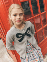 Load image into Gallery viewer, A young girl with blonde hair stands in front of a red telephone booth, wearing a gray knit cardigan with a large black bow motif on the chest, paired with a plaid skirt.