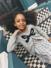 Load image into Gallery viewer, A young girl with voluminous dark curly hair smiles while lying on a black and white checkered tiled staircase. She is wearing a light gray knit cardigan with a large black bow motif on the chest, layered over a plaid skirt.