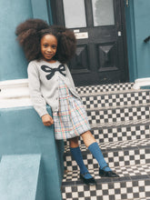 Load image into Gallery viewer, A young girl with a large afro hairstyle stands on black-and-white checkered steps in front of a teal building, wearing a light gray cardigan with a black bow, a plaid skirt, blue knee-high socks, and black shoes.