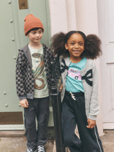 Load image into Gallery viewer, Two children standing outdoors in front of a pale green door and a light pink wall. The child on the left wears an orange beanie, a gray-and-black checkered zip hoodie over a graphic T-shirt, and dark pants.