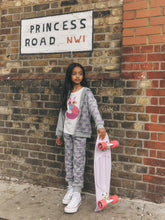 Load image into Gallery viewer, A young girl stands against a brick wall holding a pink cruiser skateboard. She is wearing a matching gray and purple floral print lounge set with a white graphic t-shirt.