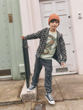 Load image into Gallery viewer, A child wearing a black and gray checkered zip-up hoodie, a green graphic t-shirt, dark gray pants, and an orange beanie stands on a sidewalk in front of a pale pink door, holding a black umbrella.