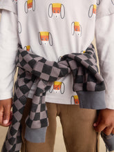 Load image into Gallery viewer, A close-up of a child wearing a white long-sleeve shirt with a repeating cartoon dog face pattern, brown pants, and a gray-and-black checkered scarf tied around the waist.