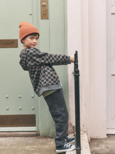 Load image into Gallery viewer, A child wearing a black and gray checkerboard zip-up hoodie, dark gray pants, and an orange beanie hat leans against a black metal post next to a pale green door.