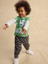 Load image into Gallery viewer, A toddler with curly dark hair is wearing a green long-sleeve graphic shirt with a soccer ball design, a white long-sleeve layer underneath, gray and black checkered pants, and white sneakers, posed against a warm beige studio backdrop.
