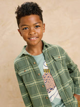 Load image into Gallery viewer, A young boy with curly dark hair smiling at the camera, wearing a green and yellow plaid button-up shirt over a light blue graphic t-shirt, set against a warm beige textured background.