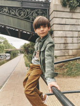 Load image into Gallery viewer, A young boy wearing a green plaid flannel shirt and brown corduroy trousers, leaning on a black metal railing next to a canal under a stone bridge.