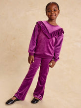 Load image into Gallery viewer, A young girl modeling a matching purple velvet two-piece outfit consisting of a long-sleeve top with a ruffled shoulder detail and matching wide-leg pants, set against a warm beige textured background.