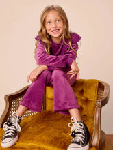 Load image into Gallery viewer, A young girl with blonde hair sits smiling on a mustard yellow velvet armchair, wearing a matching purple long-sleeve top and wide-leg pants with black high-top sneakers.