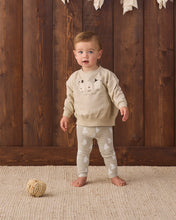 Load image into Gallery viewer, A toddler stands barefoot on a textured beige rug in front of a dark wood plank wall, wearing a cream sweatshirt with a sheep graphic and matching cream leggings with white sheep prints. A small ball of yarn sits on the rug to the left.