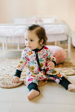 Load image into Gallery viewer, A toddler sitting on a woven rug in a softly lit bedroom, wearing a long-sleeve one-piece pajama with a colorful cartoon animal print and black trim.