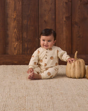 Load image into Gallery viewer, A baby sitting on a textured cream rug in front of a dark wood plank wall, wearing a cream long-sleeve romper with a brown pumpkin print. The baby is holding a small decorative pumpkin.