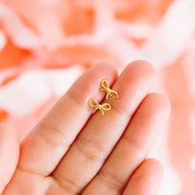 Load image into Gallery viewer, Two gold-tone bow stud earrings resting on fingertips against a soft pink background, showing their small size.
