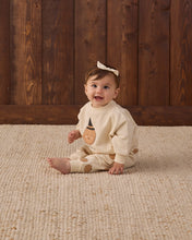 Load image into Gallery viewer, A baby sitting on a textured beige rug in front of a dark wood plank wall, wearing a cream-colored long-sleeve romper with a brown bear face graphic on the chest and matching brown bear prints on the legs, along with a cream bow headband.