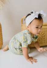 Load image into Gallery viewer, A baby crawling on a white floor wearing a pale green and yellow floral print romper and a white bow headband, with a woven basket in the background.