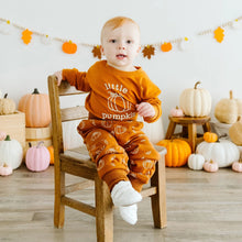 Load image into Gallery viewer, A baby sitting on a wooden chair wearing an orange long-sleeve pajama set with 'little pumpkin' and pumpkin prints, in a decorated autumn studio setting with pumpkins and fall garlands.
