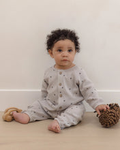 Load image into Gallery viewer, A baby with curly dark hair sits on a light wooden floor against a cream wall, wearing a light gray waffle-knit romper with small dark star prints and button closures. The baby is holding a brown pinecone toy.