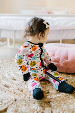 Load image into Gallery viewer, A toddler sits on a woven rug wearing a white one-piece pajama with a colorful Halloween print of pumpkins, spiders, and ghosts. The child is in a bedroom setting with a pink floor cushion and a bed in the background.