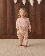 Load image into Gallery viewer, A toddler stands barefoot on a textured beige rug in front of a dark wood plank wall, wearing a matching dusty pink two-piece outfit with white polka dots. The outfit consists of a long-sleeve top and matching pants, accessorized with a white hair bow.