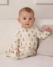 Load image into Gallery viewer, A baby sitting on a textured cream couch wearing a cream-colored footed zip-up sleeper with a repeating pattern of small brown and black illustrated characters.