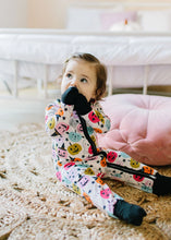 Load image into Gallery viewer, A toddler sits on a textured beige rug in a bedroom, wearing a one-piece pajama with a colorful Halloween print of jack-o'-lanterns, witches, and stars. The pajama has a white base with multicolor graphics and black accents, including a black zipper and black socks.