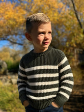 Load image into Gallery viewer, A young boy wearing a dark green and white horizontally striped knit sweater, standing outdoors in a sunny, autumnal setting with blurred yellow trees in the background.