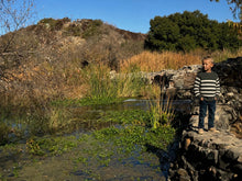 Load image into Gallery viewer, A young boy wearing a black and white striped long-sleeve shirt, dark jeans, and sneakers stands on a stone pathway next to a small pond with green aquatic plants, reeds, and a grassy hill in the background under a clear blue sky.