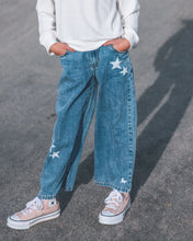Load image into Gallery viewer, A person wearing wide-leg blue denim jeans with white embroidered stars, paired with a white ribbed sweater and pink canvas sneakers, standing on a gray asphalt surface.