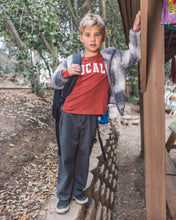 Load image into Gallery viewer, A young boy standing outdoors in a wooded area next to a wooden structure, wearing a red t-shirt with 'UCAL' printed in white, a gray patterned zip-up hoodie, dark gray sweatpants, and carrying a black backpack.