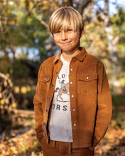 Load image into Gallery viewer, A young boy with blonde hair stands outdoors wearing a brown corduroy jacket over a white graphic t-shirt and matching brown pants, with a blurred background of autumn trees and fallen leaves.