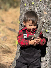 Load image into Gallery viewer, A toddler wearing a red and black plaid flannel shirt and dark overalls stands against a tree trunk outdoors, holding a small branch.