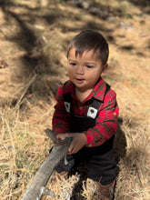 Load image into Gallery viewer, A toddler boy wearing a red and black plaid flannel shirt and black overalls sits outdoors in dry grass, holding a small branch.