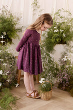 Load image into Gallery viewer, A young girl with long light brown hair wearing a long-sleeved purple dress with a small white floral pattern and brown sandals, standing in a garden-themed studio setting with potted plants and a basket.