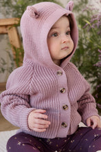 Load image into Gallery viewer, A baby wearing a dusty pink hand-knit hooded cardigan with bear ears on the hood and brown buttons, paired with dark purple star-patterned leggings, in a softly blurred garden setting.