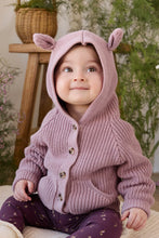 Load image into Gallery viewer, A baby wearing a chunky knit hooded cardigan with animal-ear details and button closure, paired with purple patterned leggings, sitting in a cozy indoor setting with plants and a wooden stool in the background.