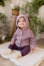 Load image into Gallery viewer, A baby sitting on a cream textured blanket in a cozy indoor setting with plants and a wooden stool. The baby is wearing a dusty mauve knit hooded cardigan with bear ears, dark purple patterned leggings, and cream booties.