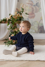Load image into Gallery viewer, A baby sitting on a cream textured rug in a styled room, wearing a navy blue cable-knit cardigan, white tights, and white shoes. A floral arrangement is on a table behind them, and a floral wall mural is on the wall.