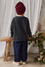 Load image into Gallery viewer, A child seen from behind wearing a dark charcoal gray knit sweater with cable-knit sleeves, navy blue pants, and a burgundy beanie, standing next to a wooden bench with flowers.