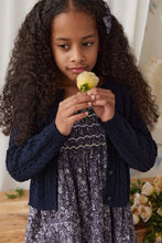 Load image into Gallery viewer, A young girl with long, curly dark hair looks down at a pale yellow rose she is holding. She is wearing a navy blue cable-knit cardigan over a purple and white floral print dress, set against a soft-focus indoor background with warm lighting.