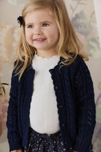 Load image into Gallery viewer, A young girl with blonde hair wearing a navy blue cable-knit cardigan over a white top and a navy patterned skirt, smiling in front of a soft floral background.