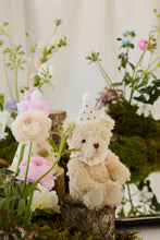 Load image into Gallery viewer, Party Arthur teddy bear seated on a wooden stump among pink flowers and moss, wearing its party hat and cream bow.