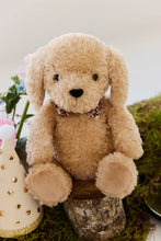 Load image into Gallery viewer, Honey the Puppy plush seated on a wooden stump, showing its floral bow tie and tan paw pads beside a gold-dotted party hat.