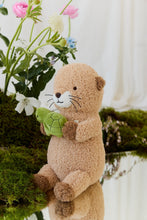 Load image into Gallery viewer, Side view of Ollie the Otter plush holding a green turtle, displayed beside moss and flowers on a mirrored surface.